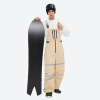 
                  
                    Unisex snowboard bib pants - tan bib pants with white top, on-model front with upright snowboard, for resort and street wear
                  
                