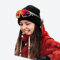 
                  
                    Warm wool knit beanie for winter sports - black wool snow beanie on women with goggles and red jacket, worn front view
                  
                